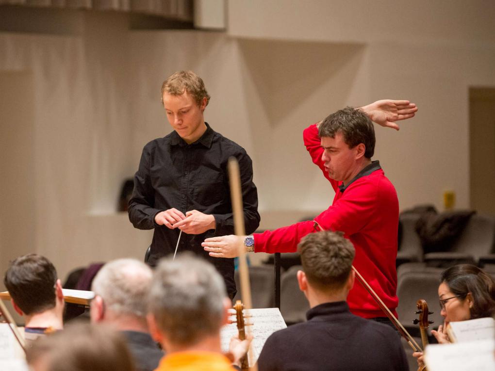 Dozent erkärt einem jungen Dirigenten etwas am Dirigierpult vor Orchester