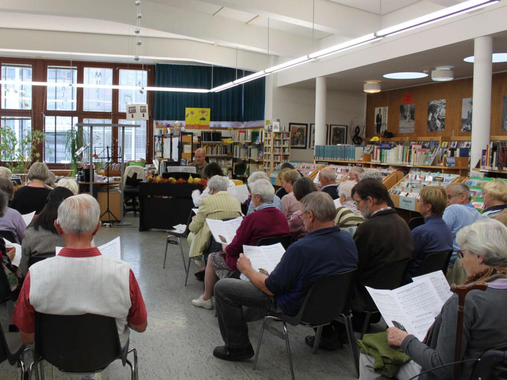 Ältere Personen singen in einem Bibliotheksraum, in der Mitte ein Herr am Klavier