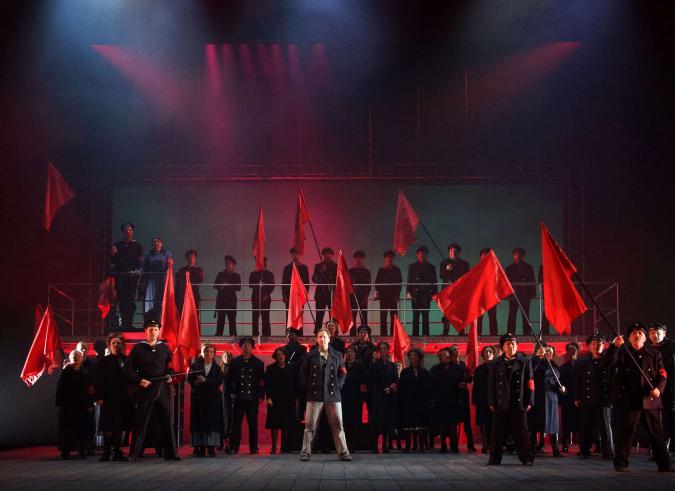 Szene auf einer Theaterbühne: Gruppe von Uniformierten mit roten Flaggen und Armbinden