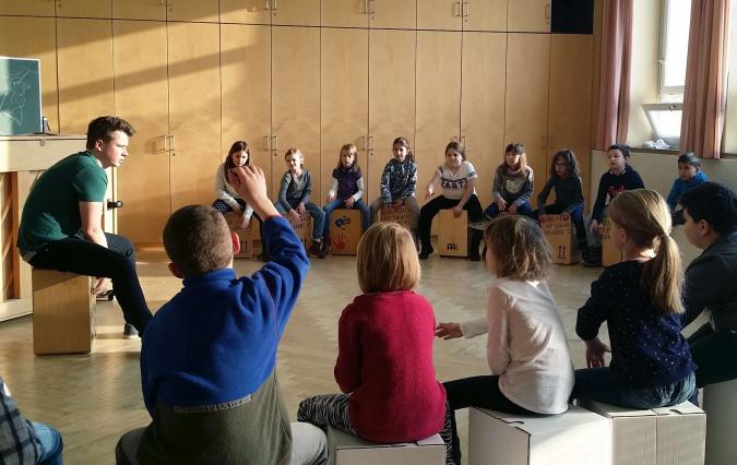 Kooperationspreojekt "Mit Musik Grenzen überwinden und Verbindung stiften": Kinder und Lehrer sitzten im Kreis und machen Musik mit Cajons.