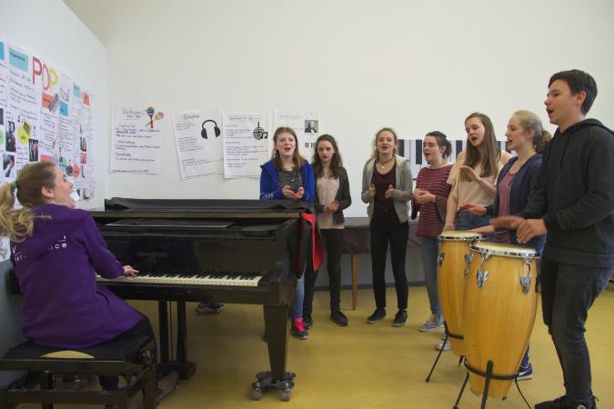 Lehrerin singt mit einer Gruppe Schülerinnen und Schülern, während sie am Klavier sitzt, die Jugendlichen klatschen und trommeln dazu