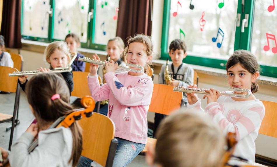 Kinder spielen gemeinsam im JeKits-Orchester auf ihren Instrumenten. Im Vordergrund des Fotos sind drei Mädchen mit ihren Kinder-Querflörten zu sehen.