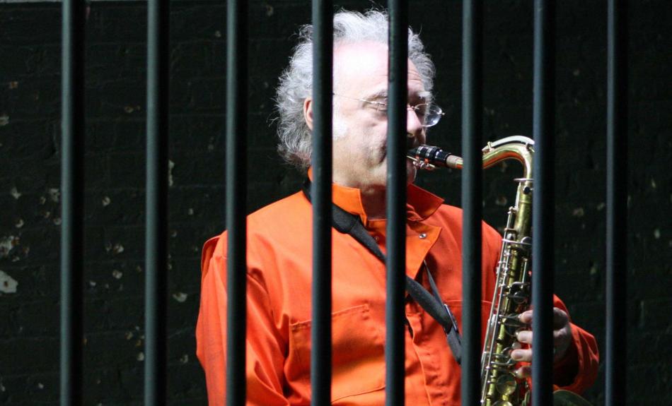 Älterer Herr in orangefarbenem Häftlingsoverall spielt Saxofon hinter Gittern