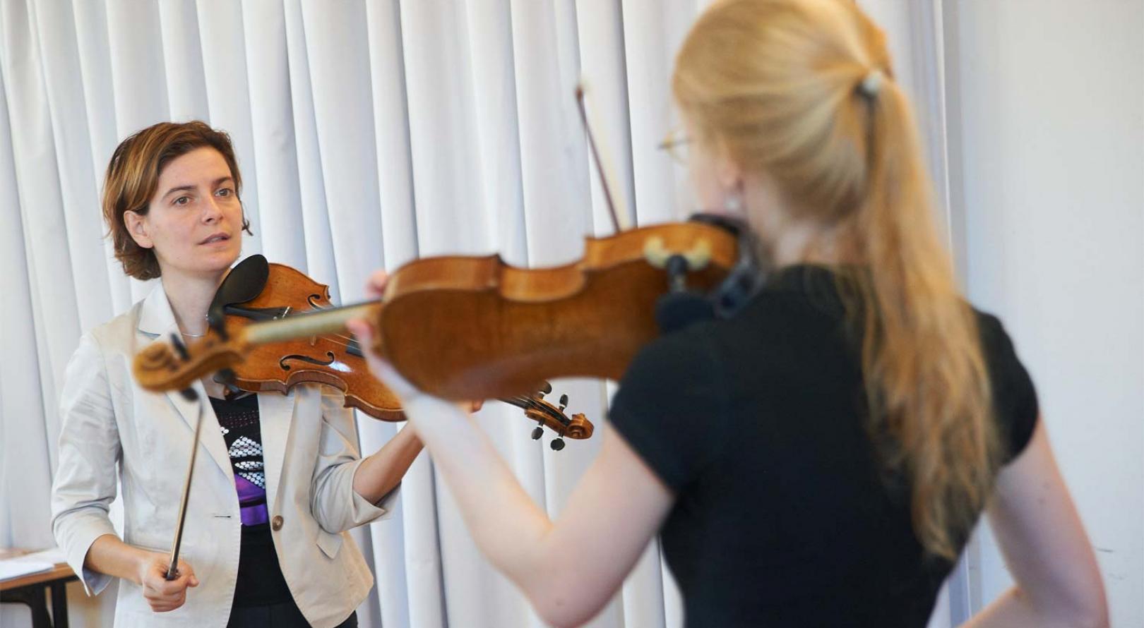 Abbildung: Teaserbild. Geigenlehrerin erklärt Studentin etwas am Instrument