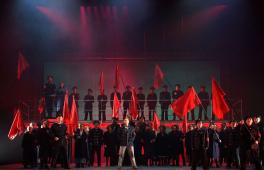 Szene auf einer Theaterbühne: Uniformierte mit roten Flaggen