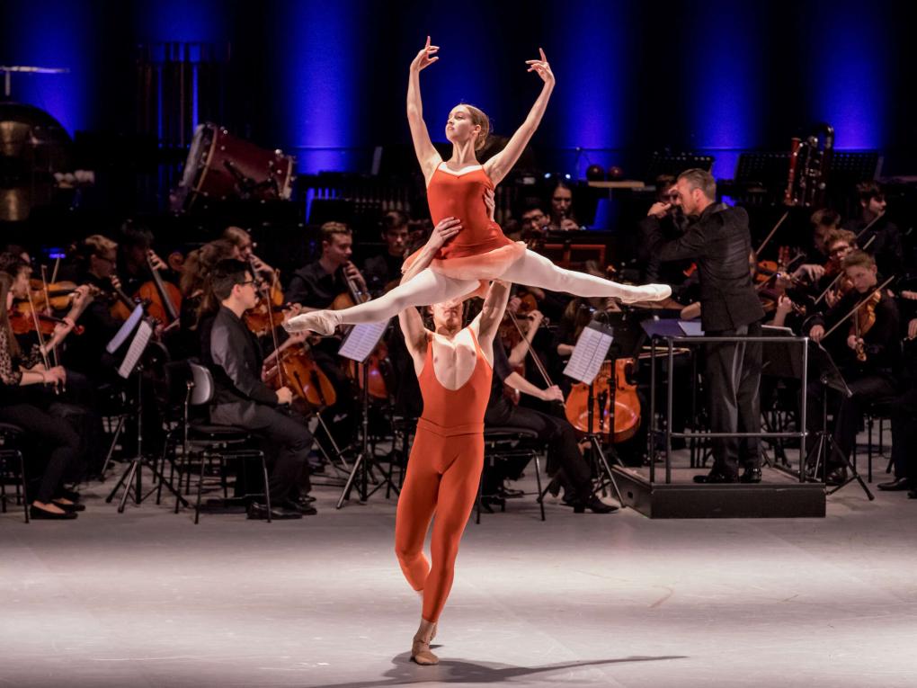 Male ballett dancer is lifting a ballerina doing the splits, an orchestra playing behind them