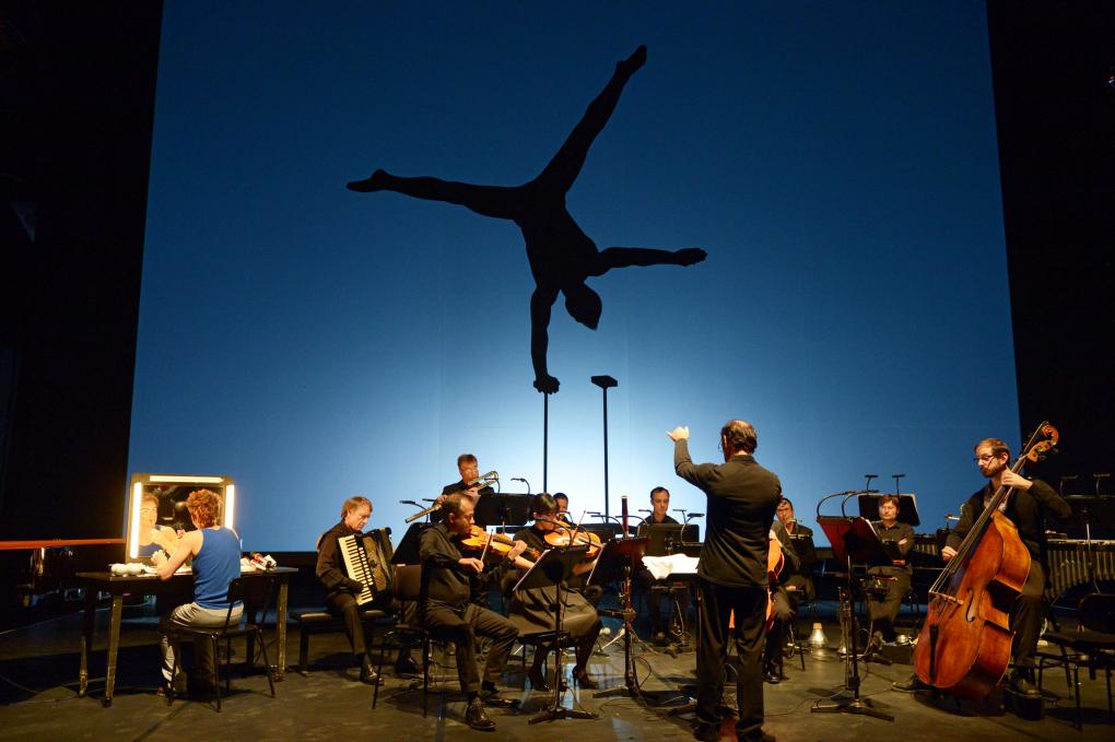 Das Ensemble Modern spielt Musik, während auf dem blauen Hintergrund die schwarze Silhouette eines Akrobaten zu sehen ist. Links auf der Bühne sitzt eine Frau vor einem beleuchteten Spiegel und trägt Make-Up auf.