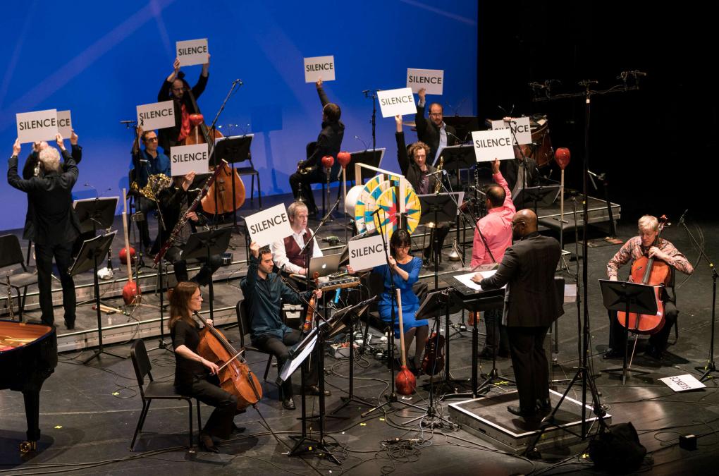 Das Ensemble Modern sitzt während der Aufführung auf Stühlen. Die Mitglieder halten Schilder mit dem Wort "Silence" über ihre Köpfe.