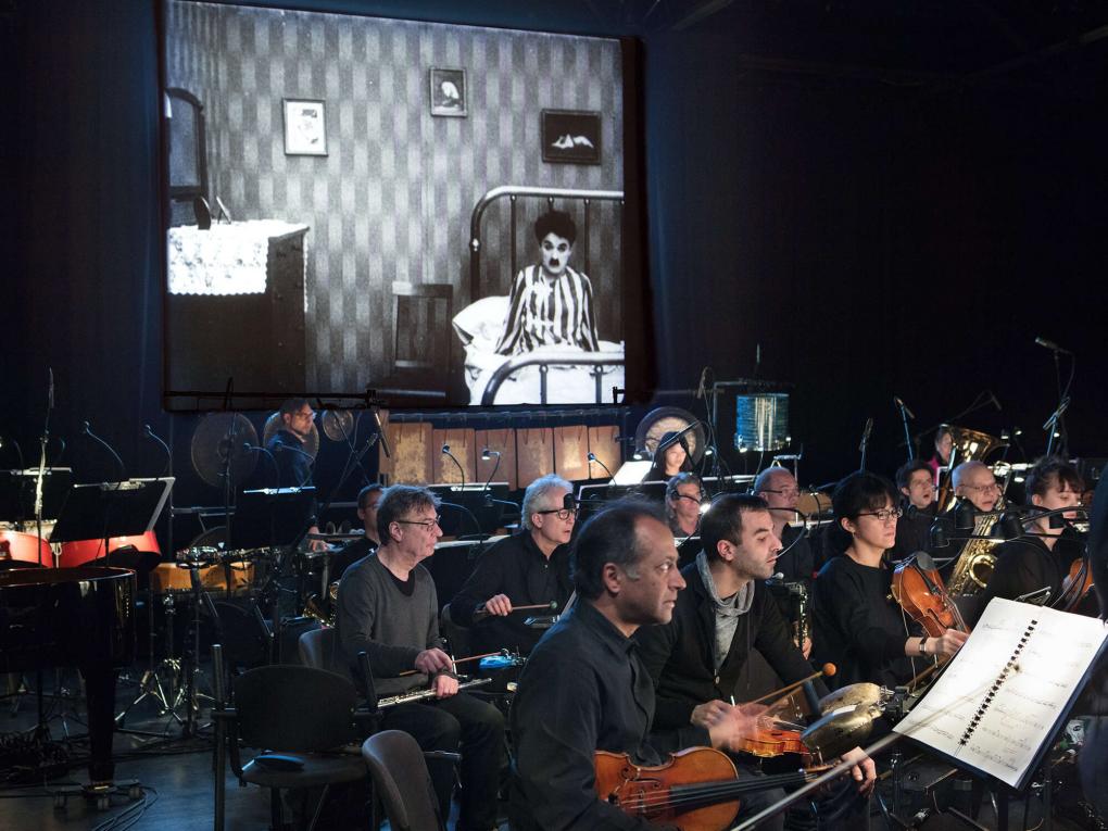 Das Ensemble Modern sitzt mit seinen Instrumenten vor einer Leinwand und wartet auf den Einsatz. Aufgeführt wird das Werk „ChaplinOperas“ von Benedict Mason. Auf der Leinwand zeigt Charlie Chaplin in einem seiner Filme.
