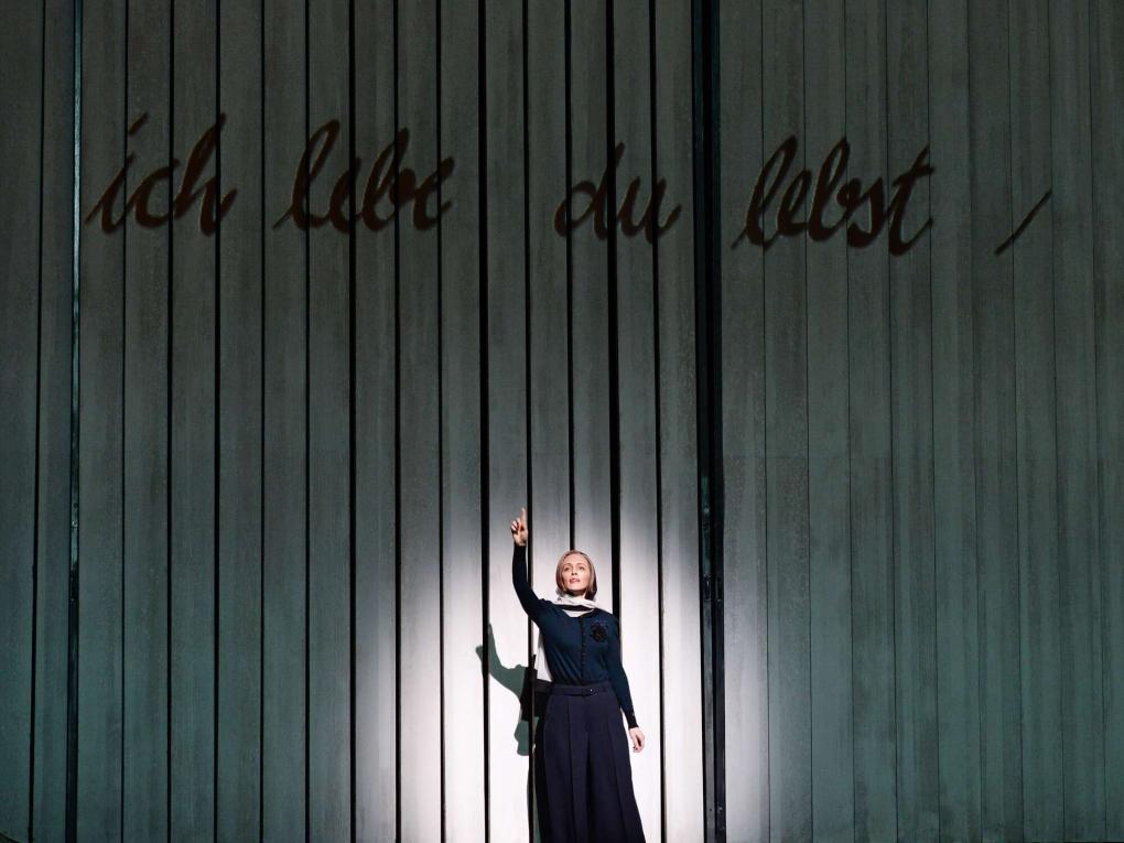 Szene auf einer Theaterbühne, Frau im schwarzen Kleid hebt den Zeigefinger vor Wand mit Aufschrift „ich lebe du lebst“