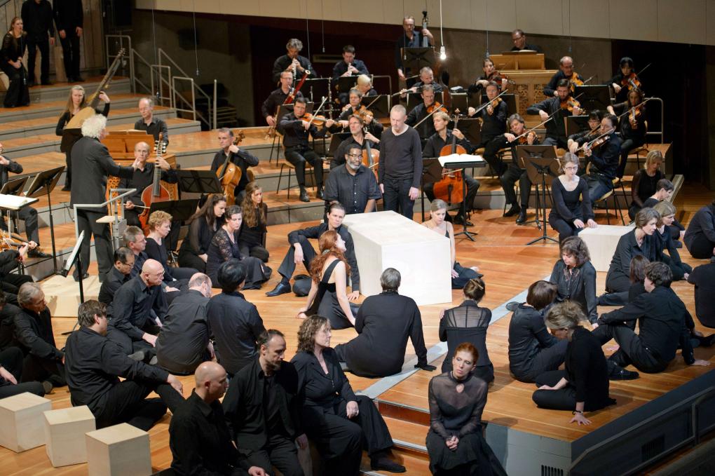 Aufführung der Matthäus-Passion in der Berliner Philharmonie, inszeniert von Peter Sellars. Die Mitwirkenden sind alle in schwarz gekleidet und sitzen über die ganze Bühne verteilt auf dem Boden. im Hintergrund sind Musikerinnen und Musiker der Berliner Philharmoniker zu erkennen, ebenfalls in schwarz gekleidet.