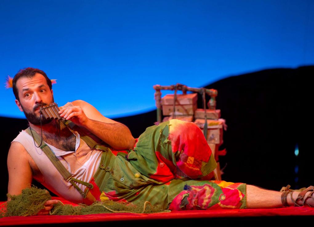 Szene auf einer Theaterbühne: liegende Person mit bunter Hose und Panflöte