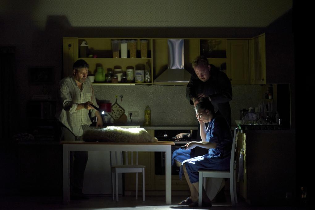 Szene auf einer Theaterbühne: Totes Lamm liegt auf Küchentisch, drei Menschen mit Taschenlampe darum herum