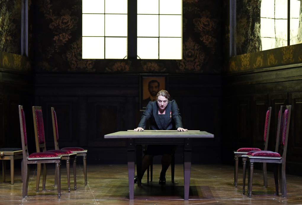 Szene auf einer Theaterbühne: Schwarz gekleidete Dame sitzt an Tisch