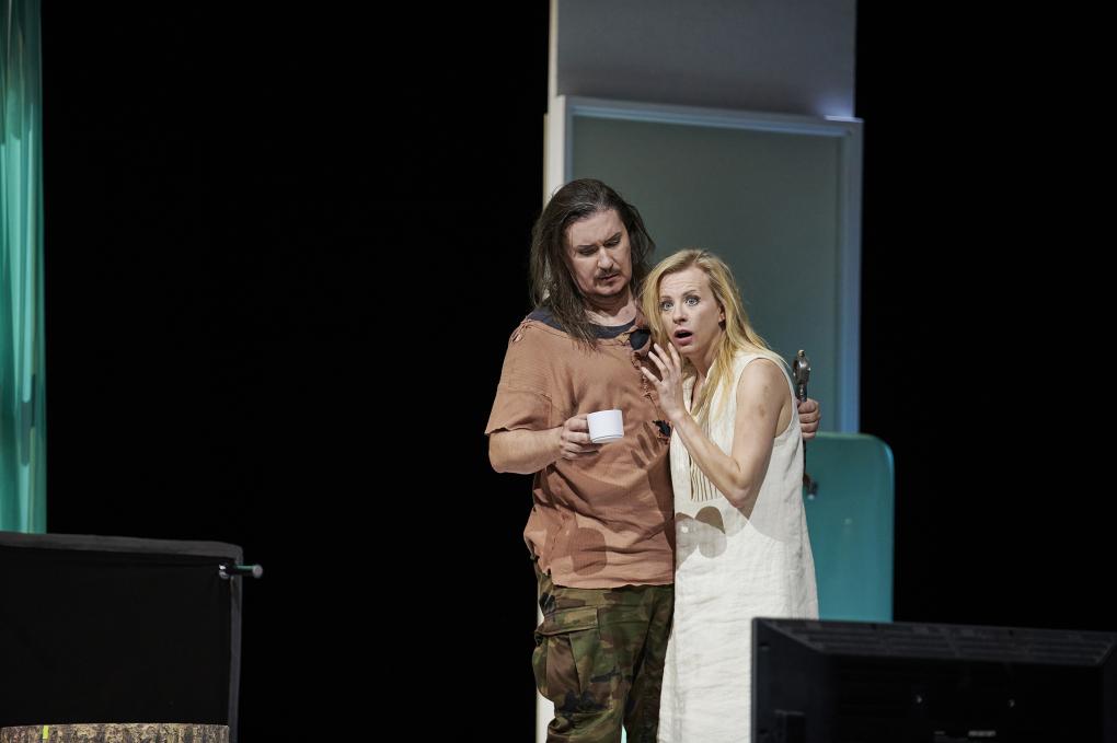 Szene auf einer Theaterbühne: Mann mit Kaffeetasse neben weißgekleideter Frau