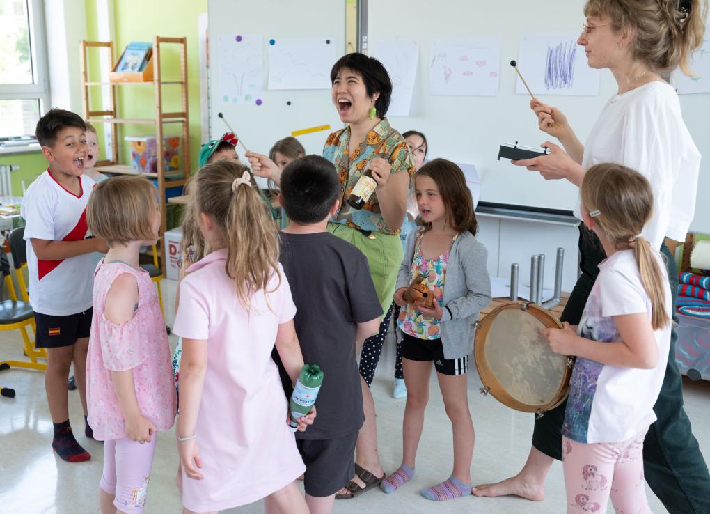 Zwei Frauen mit einer Gruppe Grundschulkinder spielen Perkussionsinstrumente.
