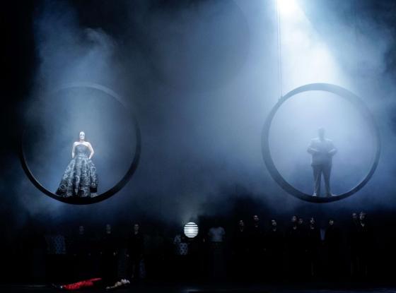 Eine Szene auf einer Opernbühne mit zwei Figuren, die in großen Ringen über der Bühne schweben